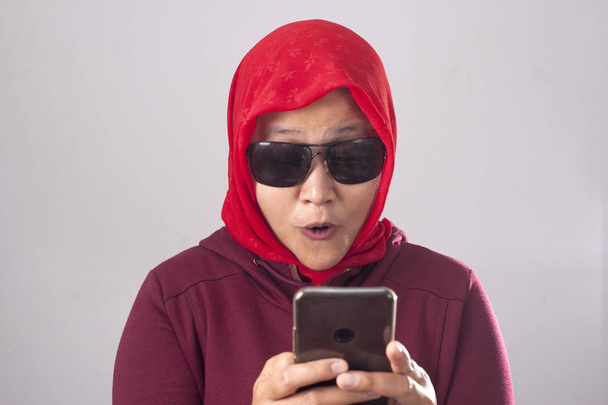 Portrait of young Asian muslim woman wearing black sunglasses in red suit and hijab get good news on her phone, happy surprised expression - Foto, Imagem