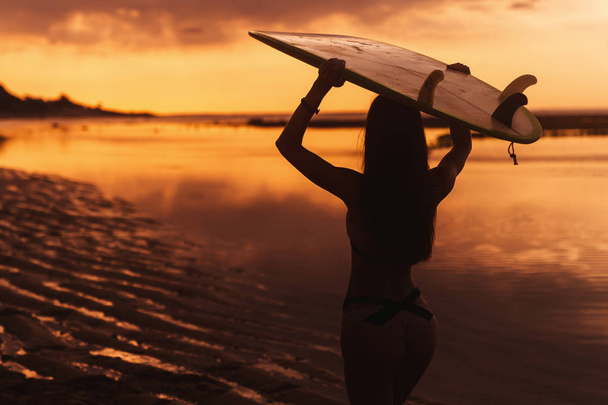 Silhouette of slim girl with surfboard in hands at beach on background of beautiful sunset. Woman go to surfing. Concept of active lifestyle, sport - Fotografie, Obrázek
