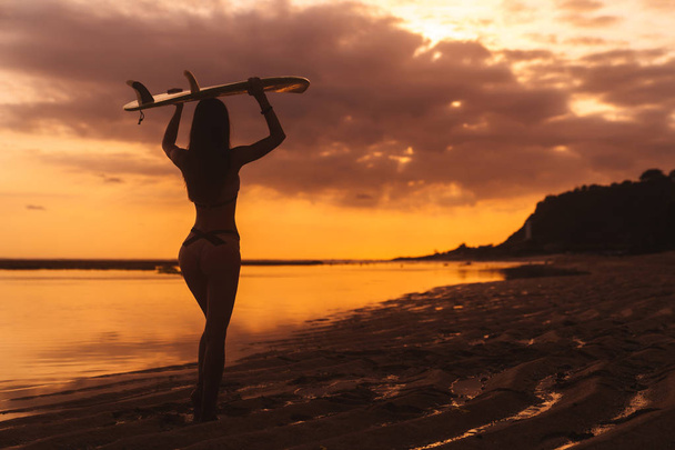 Silhouette of slim girl with surfboard in hands at beach on background of beautiful sunset - Фото, изображение