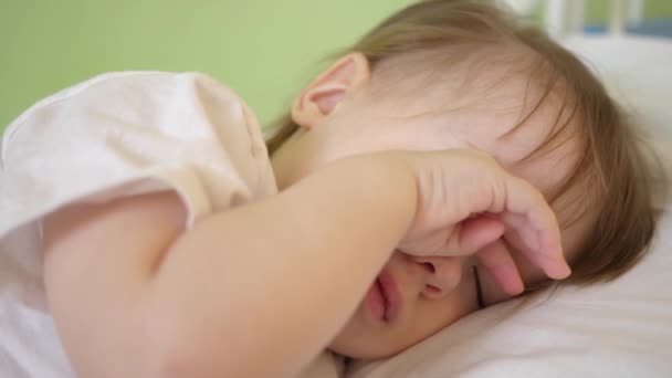 Okouzlující dítě usne na bílé posteli ve své posteli v pokoji doma. koncept spící dítě. dítě chce spát a tře si oči rukama. dítě spí v nemocniční oddělení. detail - Záběry, video