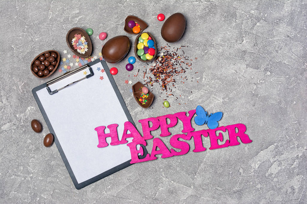 Top view on mockup of blank white sheet of paper on clipboard with pink text of happy easter and chocolate traditional eggs on gray concrete background - Fotografie, Obrázek