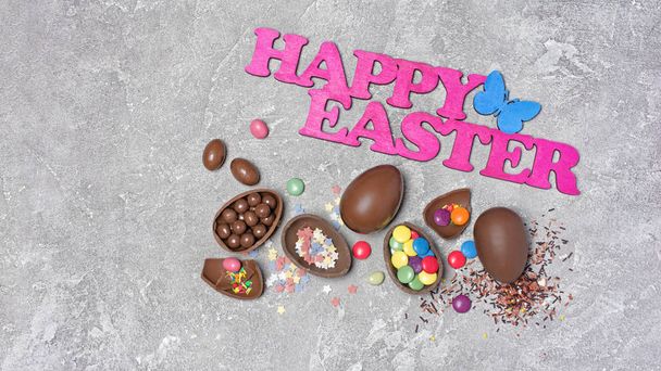 Top view on pink text of happy easter and chocolate traditional eggs with bright colorful dragee and sugar sprinkles or confetti on gray concrete background - Photo, image