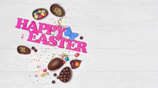 Top view on pink text of happy easter and chocolate traditional eggs with bright colorful dragee and sugar sprinkles or confetti on white wooden background with copy space - Fotografie, Obrázek