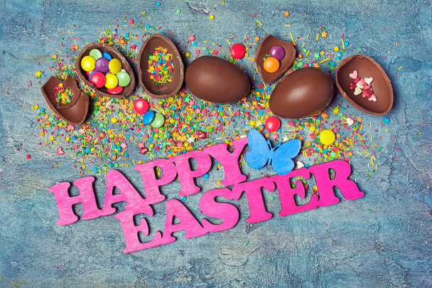 Top view on pink text of happy easter and chocolate traditional eggs with bright colorful dragee and sugar sprinkles or confetti on blue concrete background - Photo, image
