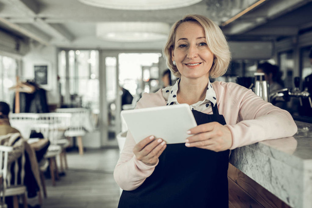 Blonde-haired entrepreneur owing restaurant holding little white tablet - Foto, Bild