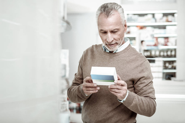 Bearded mature man reading contents of pills while buying medicine for his wife - Фото, изображение