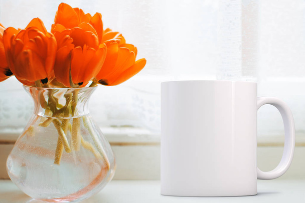 Blank mug next to a bunch of orange tulips in a glass vase on a window ledge. Perfect for businesses selling mugs, just overlay your quote or design on to the image. - Photo, Image