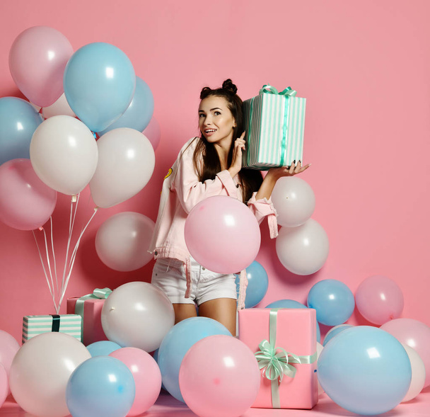 Portrait of young woman in birthday party holding gift box with gift present on pastel pink background with colorful air baloons. - Фото, зображення