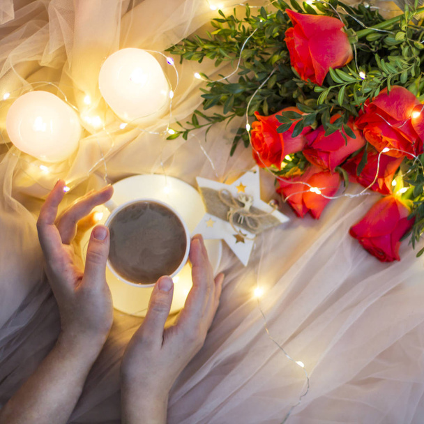 Samenstelling: rode rozen met Buxus, slingers, kaarsen, sterren en kopje koffie (thee) in de hand - Foto, afbeelding