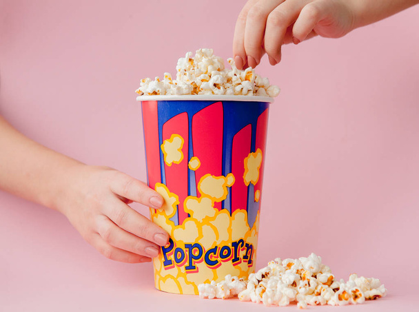 Hand takes a popcorn from a paper cup on a pink background. Woman eats popcorn.Cinema Concept. Flat lay. Copyspace. - Фото, изображение