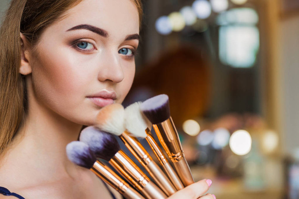 Close up the beautiful young woman holding the make-up brushes in hand near her face indoors - 写真・画像