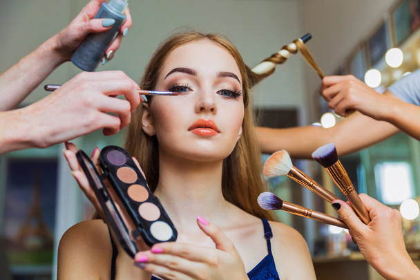 Portrait of the charming young woman who is sitting indoors and makeup artist doing makeup her and hairdresser doing the hairdo her - Foto, afbeelding
