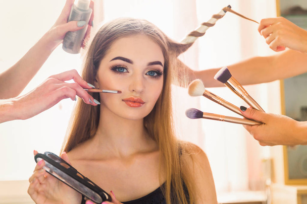 Portrait of the beautiful young woman who is sitting indoors and makeup artist doing makeup her and hairdresser doing the hairdo her - Foto, afbeelding