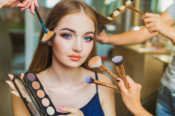 Portrait of the happy young woman who is sitting indoors and makeup artist doing makeup her and hairdresser doing the hairdo her - Foto, Bild