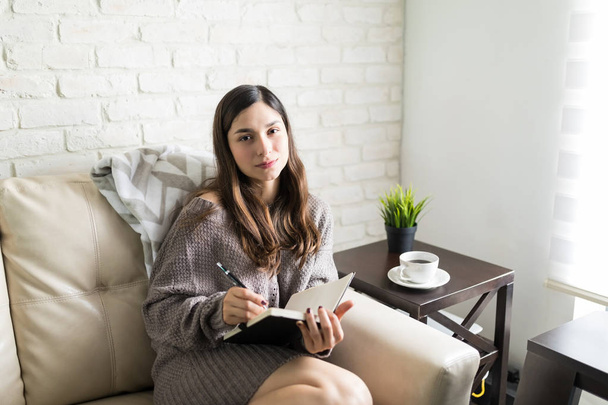 Portrait of confident woman writing notes while resting in living room - Photo, Image