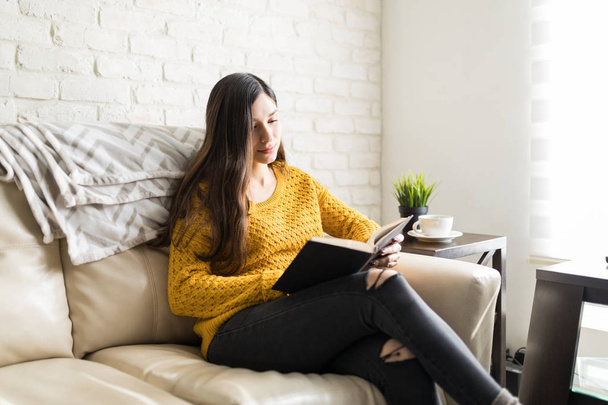 Nuori brunette nainen lukee romaania sohvalla olohuoneessa
 - Valokuva, kuva