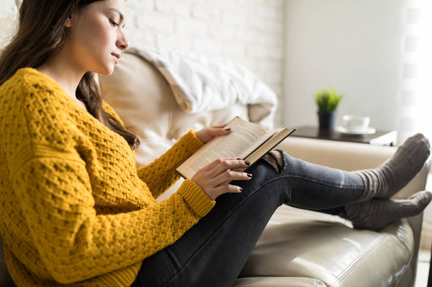 Sivunäkymä kaunis nainen keskittyy kirjan lukemiseen, kun lepää olohuoneessa
 - Valokuva, kuva