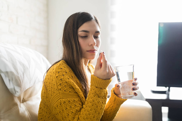 Mujer latina sosteniendo píldora y vaso de agua en la sala de estar
 - Foto, Imagen