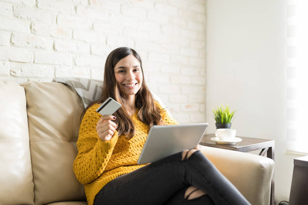 Portrait of brunette woman with plastic card and digital tablet sitting on sofa - Foto, afbeelding