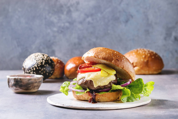 Variety of homemade classic and mini burgers in wheat and black buns with beef and veal cutlets, melted cheese and vegetables on white ceramic board over grey blue table. - Foto, Imagem