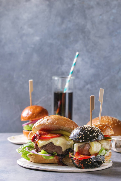 Variety of homemade classic and mini burgers in wheat and black buns with beef and veal cutlets, melted cheese and vegetables on white ceramic board over grey blue table. With glass of cola. - Foto, Imagem