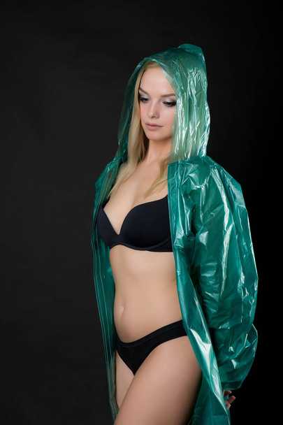 Portrait of a young beautiful blond woman in a green raincoat looking up, a hood is put on her head. Shooting in a photo studio on a black background - Foto, imagen
