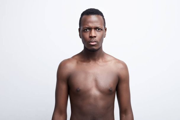 Close up shot of serious dark skinned male with scared and anxious emotion on face, feels astonishment as sees something unexpected, posing shirtless, isolated over white background. Human expressions - 写真・画像
