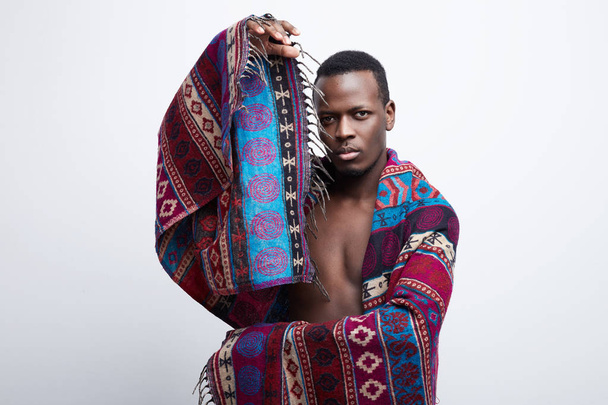 Authentic portrait of handsome dark skin brutal male, standing isolated on white studio background, posing alone, wearing ethnic colorful blanket and playing with it, sensually looking at camera. - Photo, image