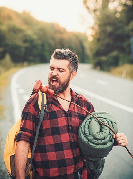 Man hiker eat sausage on stick. Bearded man with food hiking on road. Hipster with backpack and sleeping bag. Traveler travel and hiking on summer vacation. Eating food concept - Фото, изображение