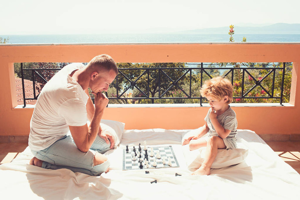 father with son on balcony play chess. Child play chess with father. Love and trust as family values. Family travel with kid on fathers day. Summer vacation of happy family - 写真・画像