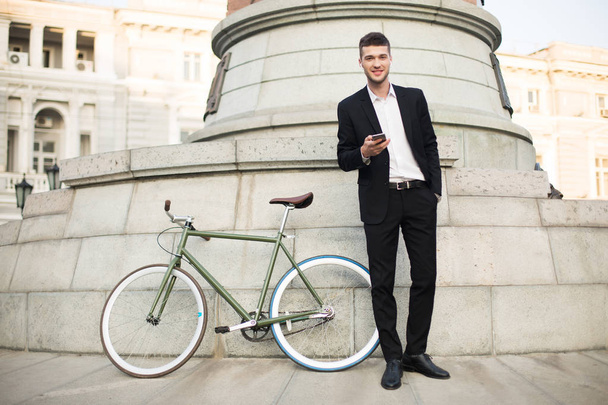Jeune homme d'affaires souriant en costume noir classique et chemise blanche avec écouteurs sans fil tenant le téléphone portable à la main regardant à la caméra avec vélo rétro près de la rue
 - Photo, image