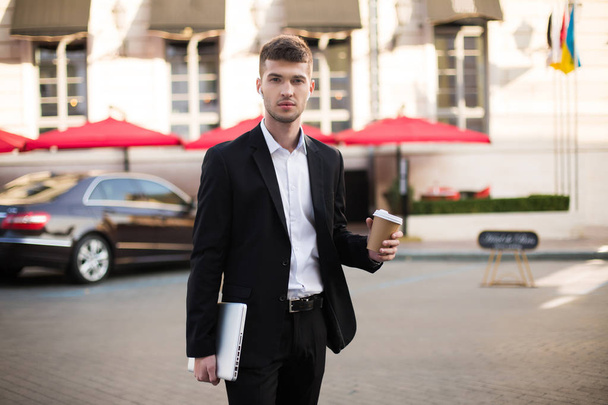 Jeune homme sérieux en costume noir et chemise blanche avec écouteurs sans fil tenant un ordinateur portable et un café dans les mains regardant attentivement à la caméra avec une belle rue sur fond
 - Photo, image