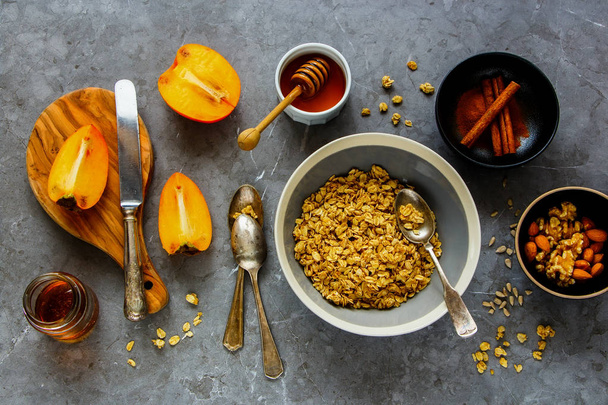 Healthy breakfast variety. Flat-lay of cinnamon granola, fresh persimmon, maple syrup, honey and nuts over grey concrete table background, top view. Clean eating, dieting, vegetarian food - Image - Photo, image