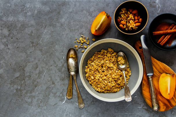 Flat-lay of cinnamon granola bowl, fresh persimmon and nuts over grey concrete table background, top view. Clean eating, dieting, vegetarian food - Image - Foto, Imagem