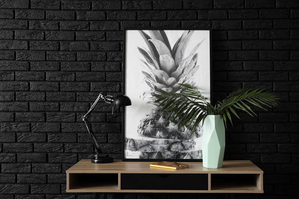 Vase avec feuilles tropicales, photo et lampe sur la table près du mur de briques sombres
 - Photo, image
