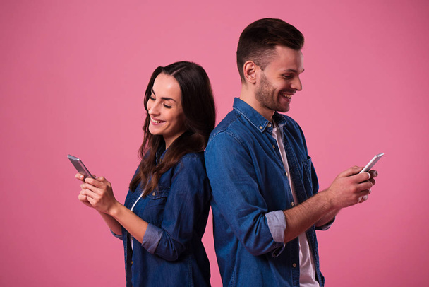 fröhliches junges Paar, isoliert vor rosa Hintergrund stehend, Smartphones in Freizeitkleidung haltend - Foto, Bild