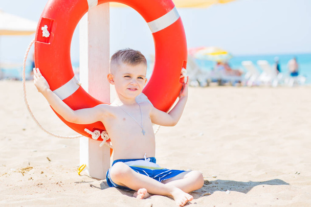 terve pieni poika istuu rannalla pelastusrenkaan kanssa kesällä aurinkoinen päivä
 - Valokuva, kuva
