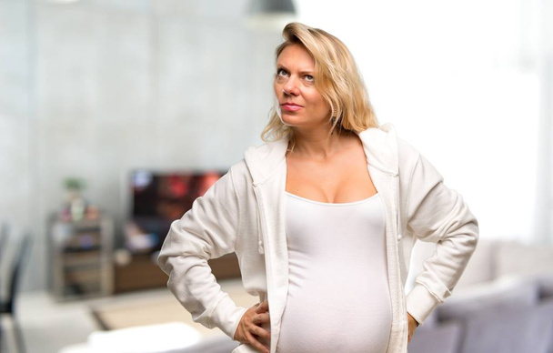 Pregnant blonde woman with white sweatshirt having doubts and with confuse face expression in her house - Fotoğraf, Görsel