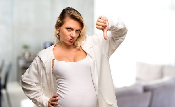 Mulher loira grávida com camisola branca mostrando sinal polegar para baixo com expressão negativa em sua casa
 - Foto, Imagem