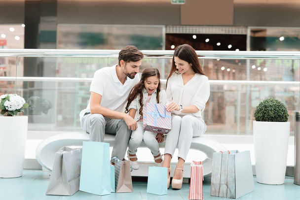 Family, father, mother and daughter are sitting on bench in shopping mall. Girl is looking inside one bag. - Zdjęcie, obraz