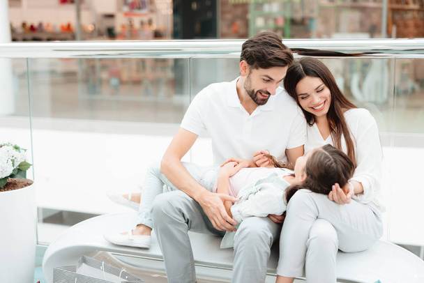 Семья из трех человек, отец, мать и дочь сидят на скамейке в торговом центре
. - Фото, изображение
