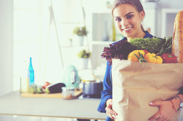 Giovane donna che tiene borsa della spesa con verdure. In piedi in cucina - Foto, immagini