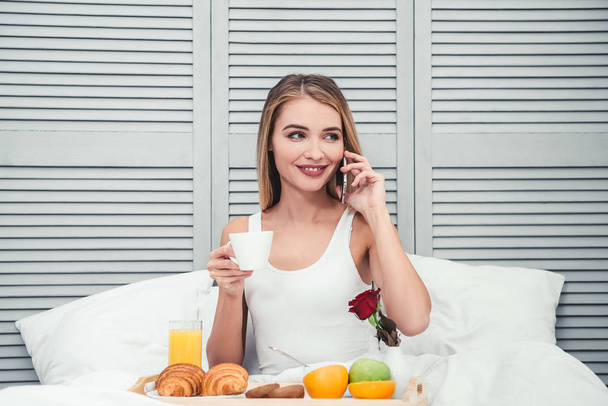 cheerful woman calling on smartphone while having breakfast in the bed at the morning - Foto, Imagen