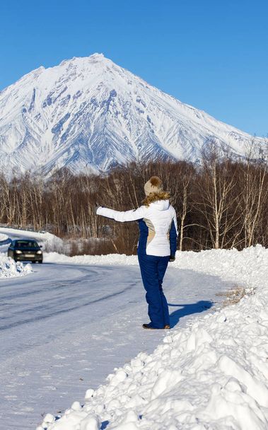 The girl hitchhiking on the winter road opposite the volcano  - Photo, Image