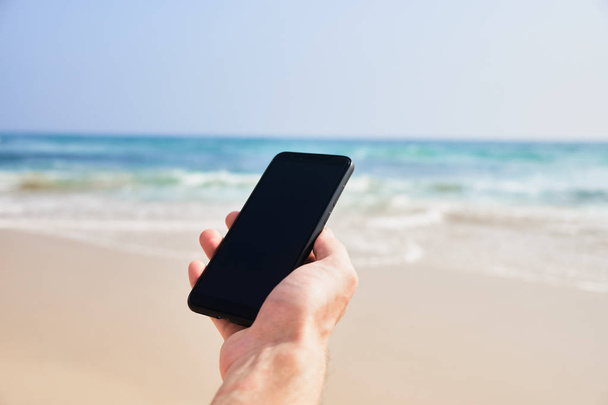 Mobile phone in male hand on the background of the beach and ocean. - Foto, Imagem