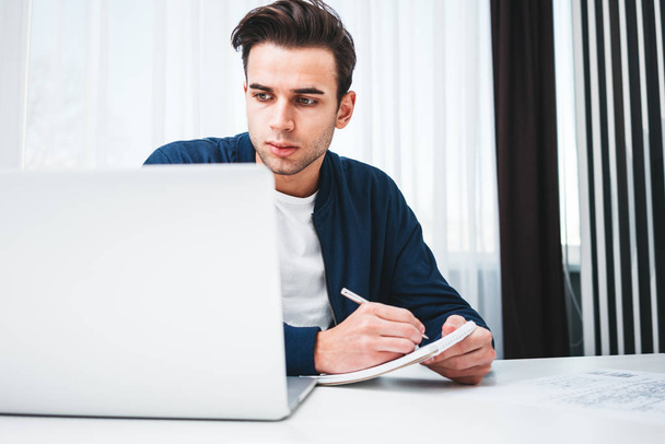 Hombre concentrado escribiendo nuevas ideas en bloc de notas mirando en la computadora portátil de pantalla. Joven compañero de trabajo se sienta a la mesa y trabaja en la oficina
 - Foto, Imagen