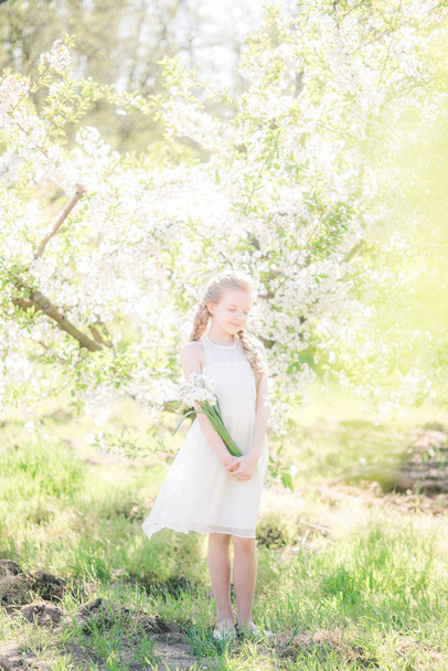 Cute girl with blond hair in a white sundress in spring in a lush garden with daffodils - Фото, изображение