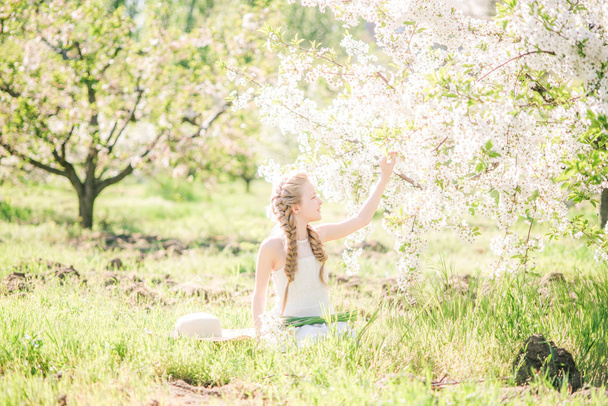 Cute girl with blond hair in a white sundress in spring in a lush garden with daffodils - Foto, immagini
