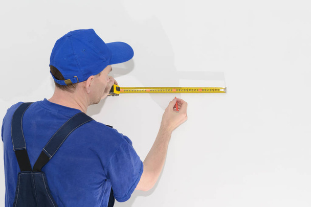 worker in blue workwear measures the distance on a white wall using a tape measure - Foto, immagini