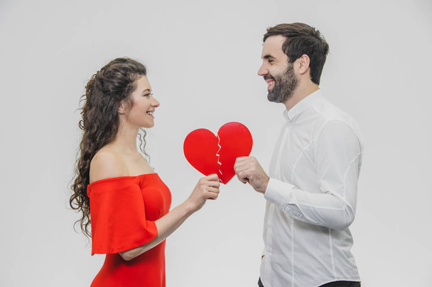 Portrait of a Loving Young Couple. A man is dressed in a white shirt, his wife in a red dress. Conducting two parts of the heart paper. - Фото, зображення